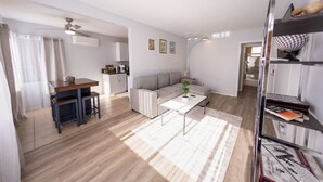 Ein geräumiger Wohnbereich mit einer Couch, einem Couchtisch und einem Esstisch. Im Hintergrund ist eine Küche zu sehen, und ein Bücherregal mit Büchern steht daneben.