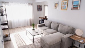 Ein modernes Wohnzimmer mit einem grauen Ecksofa, einem Couchtisch und einem kleinen Beistelltisch mit Lampe. Ein Bücherregal mit Büchern und Dekorationselementen, sowie eine kleine Topfpflanze auf dem Couchtisch.