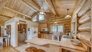 A rustic cabin-style kitchen with wooden beams, a central island with stools, a refrigerator with magnets, and a window overlooking the forest. - Inviting and Relaxing-Cliffs and Streams! (Nathrop)