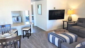 Un salon avec un canapé gris, un pouf à rayures et un téléviseur fixé au mur. On y trouve également un coin repas avec une table et des chaises, et une cuisine à l’arrière-plan.