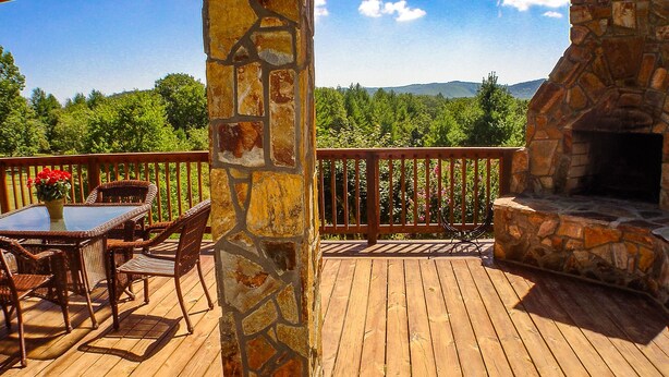 Una terraza de madera con chimenea de piedra, mobiliario de mimbre y vistas a un paisaje boscoso.