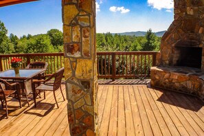 Una terraza de madera con chimenea de piedra, mobiliario de mimbre y vistas a un paisaje boscoso.