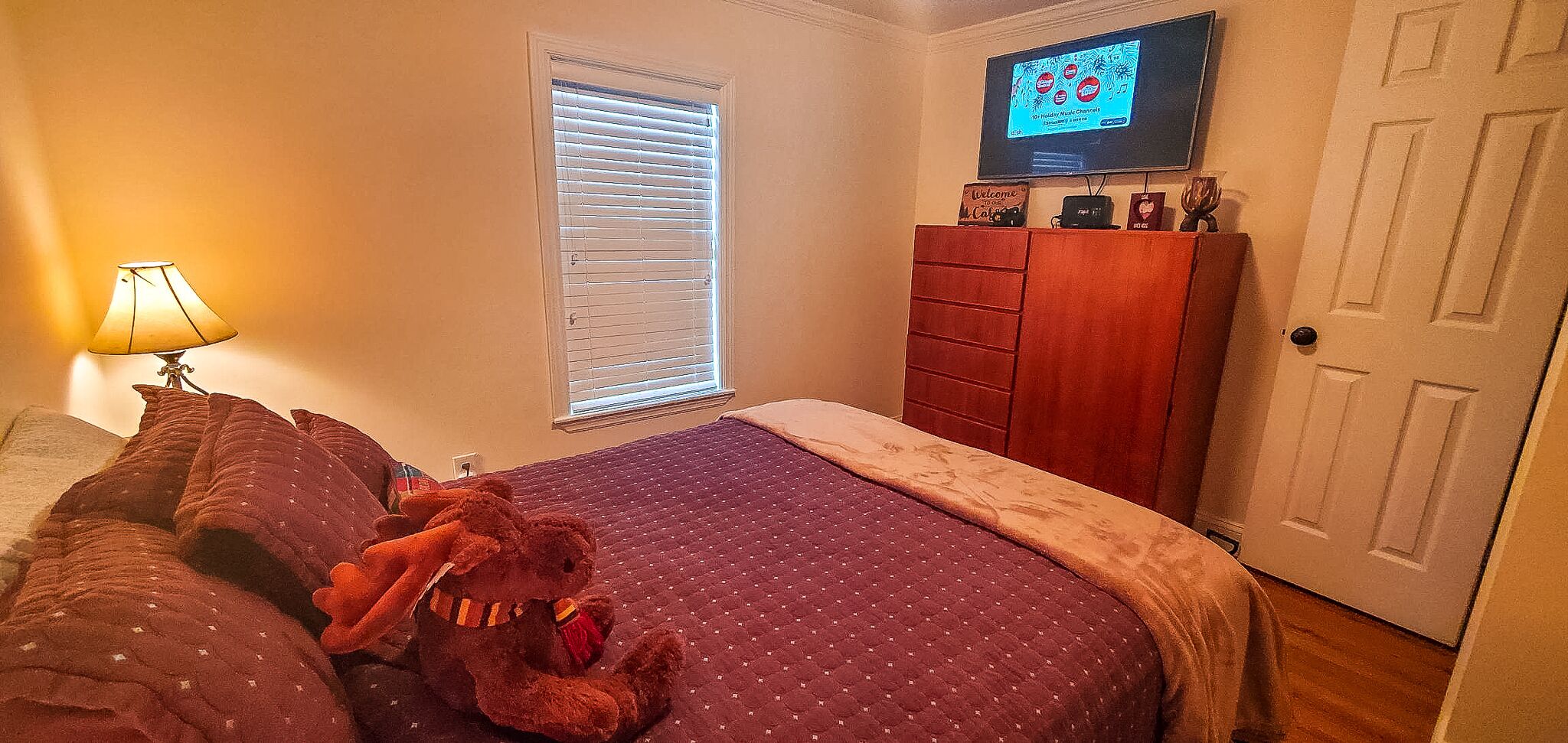 A bedroom with a bed, a window with blinds, a television on the wall, and a wooden dresser.