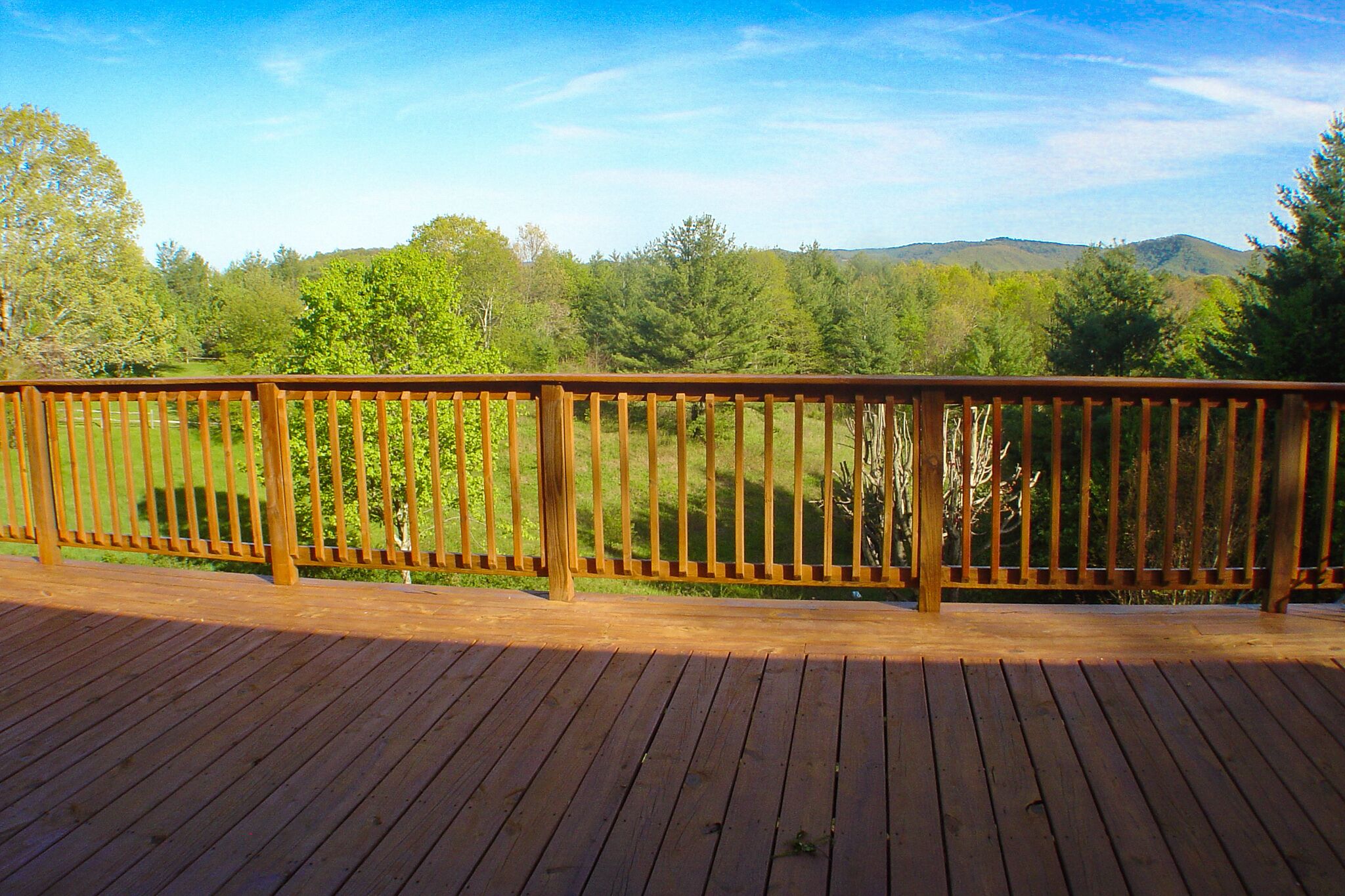 A wooden deck with a railing overlooking a grassy area with trees and hills in the background.