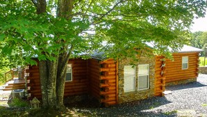 Una cabaña de troncos con cimientos de piedra, una terraza de madera y un árbol grande al frente.