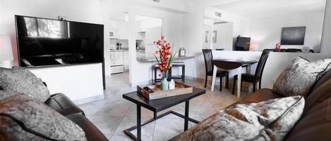 A modern living room with a flat-screen TV, a dining area with a table and chairs, and a kitchen in the background.