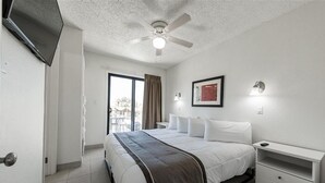 A hotel room with a bed, a ceiling fan, a nightstand, a lamp, and a view of the outdoors.
