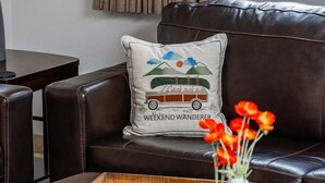 A dark brown leather sofa with a cushion featuring a car and the text 'WEEKEND WANDERER.' A wooden tray with a vase of red flowers and a small white vase is on a dark wooden coffee table. - 237- Fully Furnished with WiFi by RedAwning (Scottsdale)