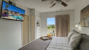 Une chambre d’hôtel équipée d’un lit, d’un ventilateur de plafond, d’un téléviseur diffusant une vue sur la piscine et d’un balcon avec une table et des chaises.