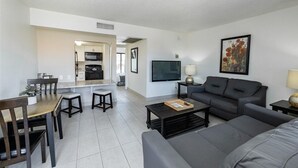 A modern living room with a grey sofa, a coffee table, a dining area with a table and chairs, a kitchen area with a microwave, and a flat-screen TV mounted on the wall.