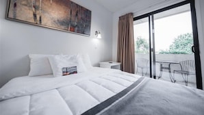 A hotel room with a bed, a painting, a nightstand, a lamp, and a balcony with chairs.