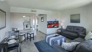 A modern living room with a grey sofa, a TV mounted on the wall, and a dining area with a table and chairs.