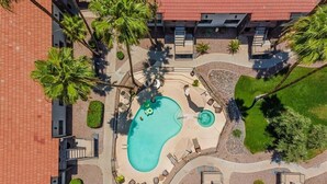 An aerial view of a hotel with a swimming pool, palm trees, and a red-roofed building.