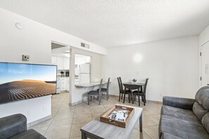 Uma sala de estar moderna com uma TV de tela plana, um sofá cinza, uma mesa de centro com revistas e uma área de jantar com mesa e cadeiras.