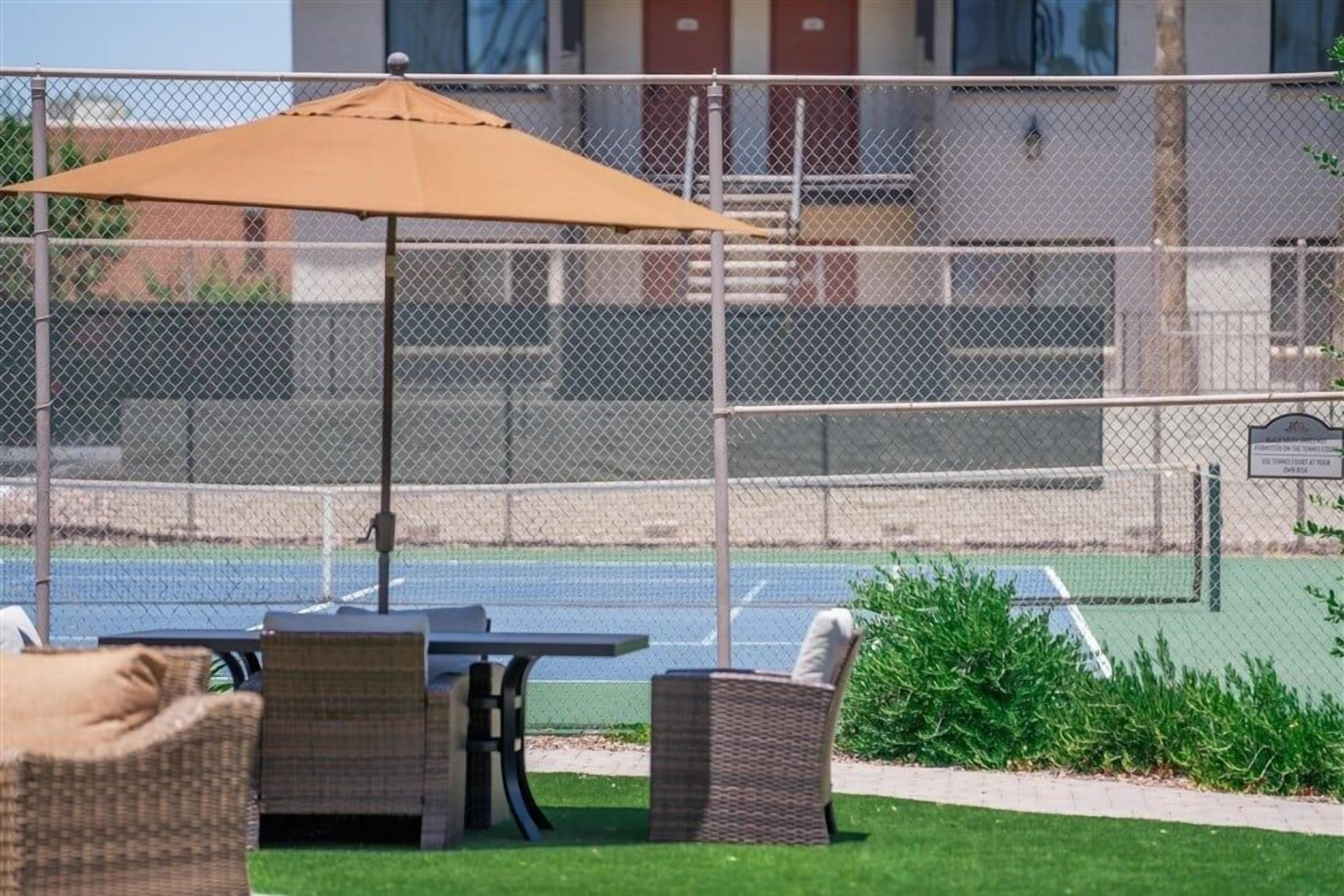An outdoor seating area with a table and chairs, a patio umbrella, and a tennis court in the background.