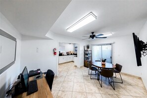 Uma cozinha moderna com armários brancos, uma mesa de jantar com cadeiras, um ventilador de teto e uma janela grande com persianas.