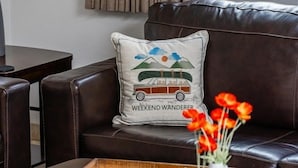 A dark brown leather sofa with a cushion featuring a car and the text 'WEEKEND WANDERER.' A wooden tray with a vase of red flowers and a small white vase is on a dark wooden coffee table. - Fully Furnished 1BR Suite-Prime Location! 110 by RedAwning (Scottsdale)