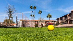 Ein grüner Rasen aus Kunstfaser mit einem gelben Ball und bunten Bällen, ein eingezäuntes Areal sowie Gebäude mit Balkonen und Palmen im Hintergrund.