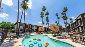 Ein Swimmingpool mit Liegestühlen und Sonnenschirmen, umgeben von Palmen und einem Gebäude mit Balkonen.