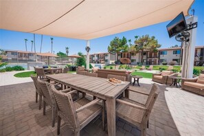 An outdoor patio area with a wooden table and wicker chairs, surrounded by a covered seating area with sofas and a TV mounted on a column.