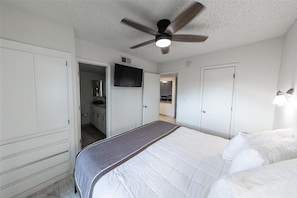 A bedroom with a bed, a ceiling fan, a TV, and a door leading to a bathroom.