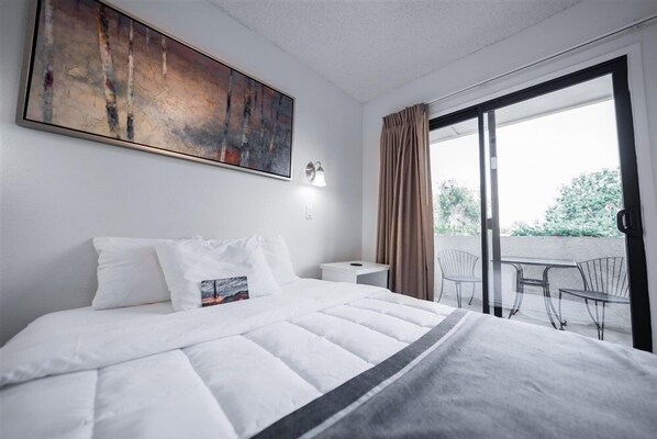 A hotel room with a bed, a painting, a nightstand, a lamp, and a balcony with chairs.