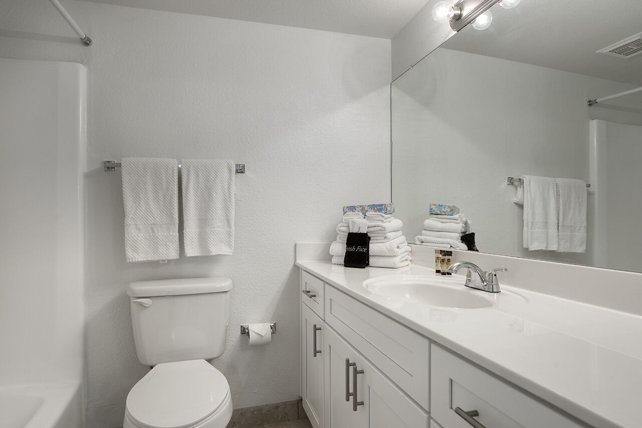A modern bathroom with a white sink, mirror, and toilet. There are towels and toiletries on the counter.
