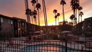 A pool area with lounge chairs and a lifebuoy, surrounded by buildings and palm trees, during sunset.