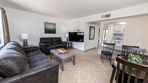 A living room with a sofa, a coffee table, a TV, and a kitchen area visible through an open doorway.