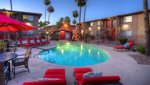 A pool area with red lounge chairs and umbrellas, surrounded by buildings with balconies and palm trees.