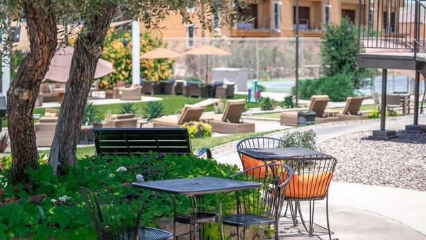 An outdoor seating area with a table and chairs, surrounded by greenery and a pool in the background.