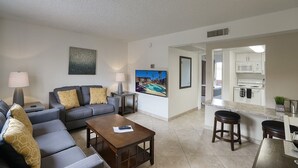 Una sala moderna con un sofá gris, una mesita de centro y un televisor. Hay una zona de cocina con un microondas y un lavabo.