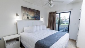 Ein Schlafzimmer mit Bett, Nachttisch, Gemälde, Deckenventilator und Blick durch eine Glastür.