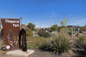 Um portão de metal com a inscrição 'Chaparral Park', cercado por diversas plantas e um céu azul claro.