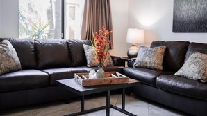 Een woonkamer met een donker leren bank, een houten salontafel met een bloevas, en een lamp op een bijzettafeltje.