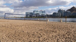 Een strandvolleybalveld met een net, een zandstrand en een stadsgezicht op de achtergrond, inclusief moderne gebouwen en een heuvel.