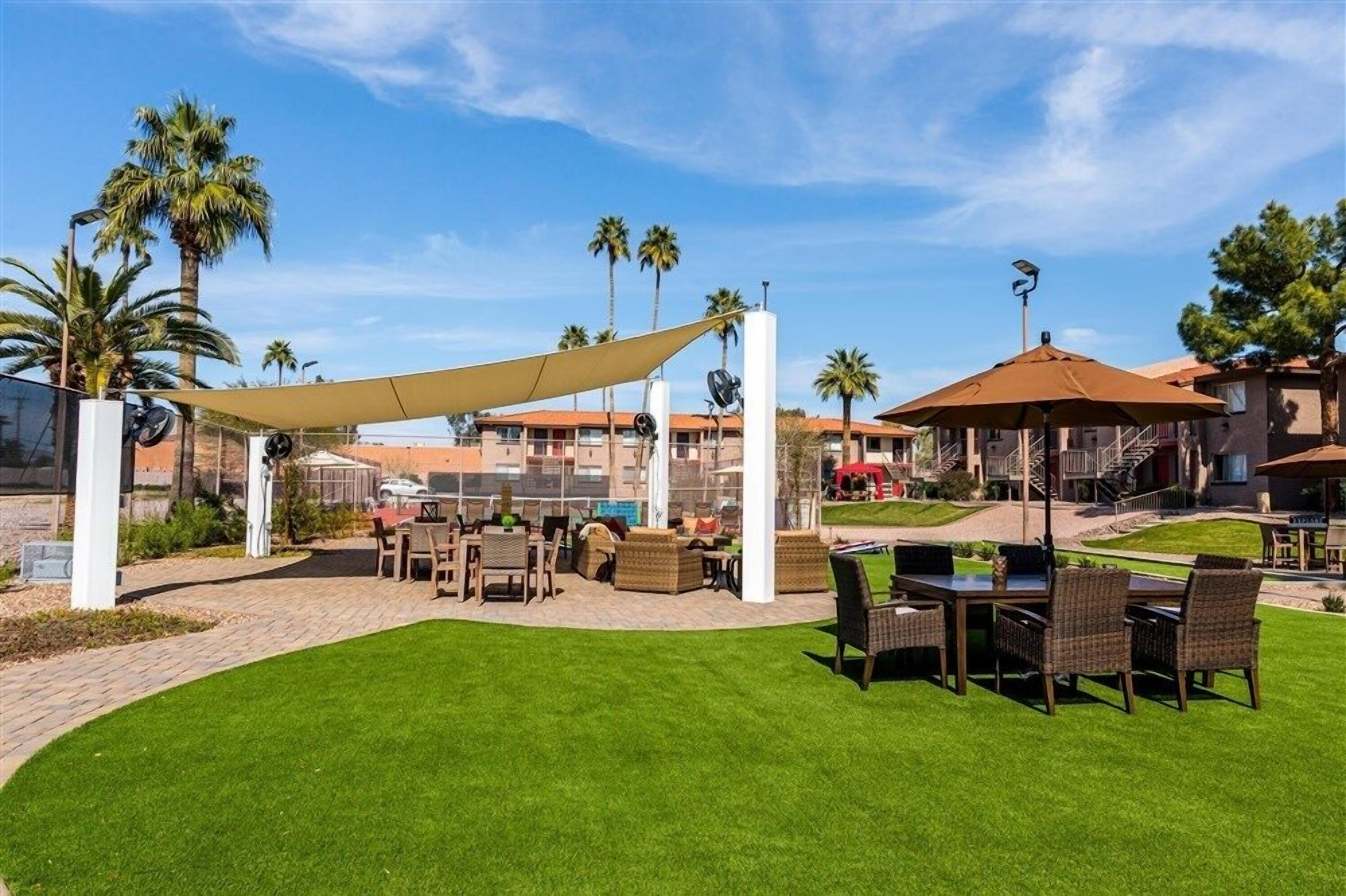 Un espace extérieur aménagé en terrasse, avec des tables et des chaises, entouré de palmiers et surplombé d’un ciel dégagé.