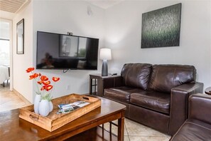 Uma sala de estar com um sofá de couro marrom, uma mesa de centro de madeira com um porta-bandejas de flores vermelhas, uma TV de tela plana, um abajur e um quadro na parede.
