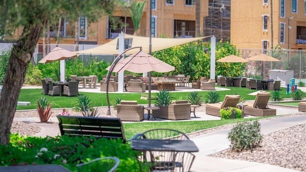 An outdoor patio area with multiple tables and chairs, shaded by umbrellas, surrounded by greenery and a building in the background. - 116 Fully Furnished 1BR Suite-Prime Location! by RedAwning (Scottsdale)