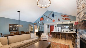 A modern living room with a sofa, coffee table, and a dining area with chairs and a table. There is a kitchen with a bar and a stone fireplace. - Fraser Crossing/Founders Pointe, Condo | 3 bedroom Ski Slope Views (Select Rated Condo 3625) (Winter Park)