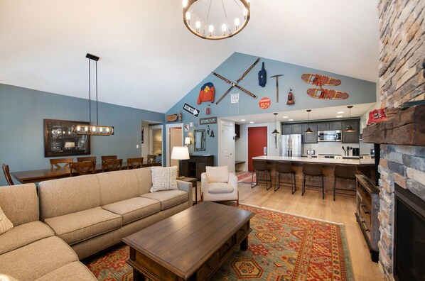 A modern living room with a sofa, coffee table, and a dining area with chairs and a table. There is a kitchen with a bar and a stone fireplace. - Fraser Crossing/Founders Pointe, Condo | 3 bedroom Ski Slope Views (Select Rated Condo 3625) (Winter Park)