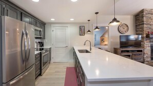 A modern kitchen with stainless steel appliances, a large island, pendant lighting, and a stone fireplace. - Fraser Crossing/Founders Pointe, Condo | 3 bedroom Ski Slope Views (Select Rated Condo 3625) (Winter Park)