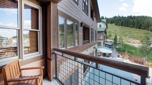 A wooden balcony with a wooden bench, large windows, and a view of a green landscape with a building in the distance. - Zephyr Mountain Lodge, Condo | 1 Bedroom - (Premium-Rated Condo 2319) (Winter Park)