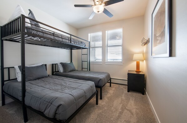 A dormitory room with bunk beds, a ceiling fan, a bedside table with a lamp, and a framed picture on the wall.