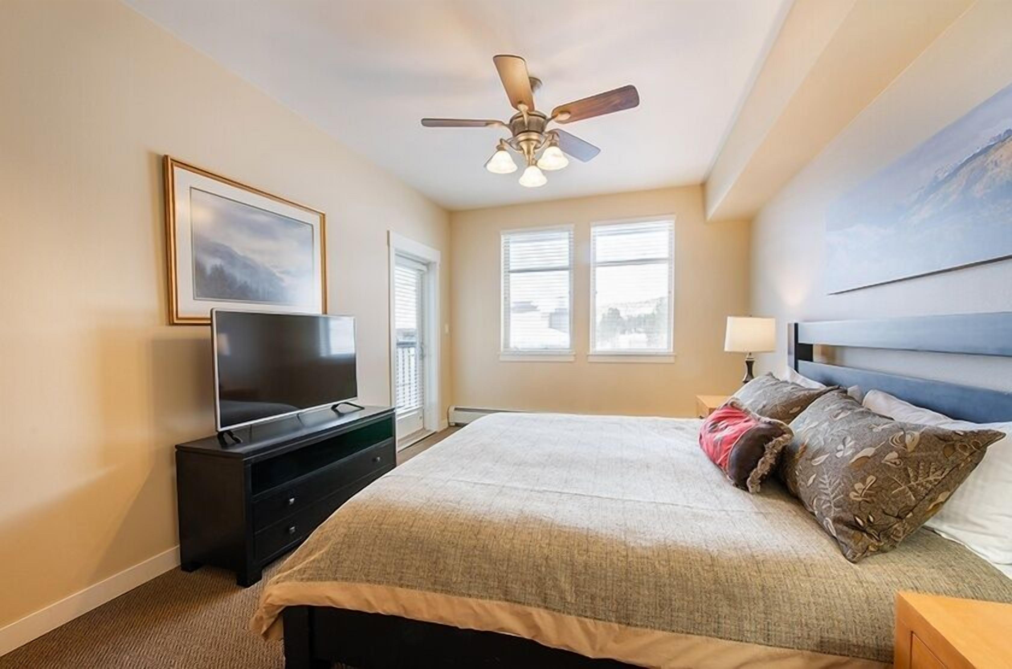A bedroom with a bed, a TV on a stand, a ceiling fan, and two windows.