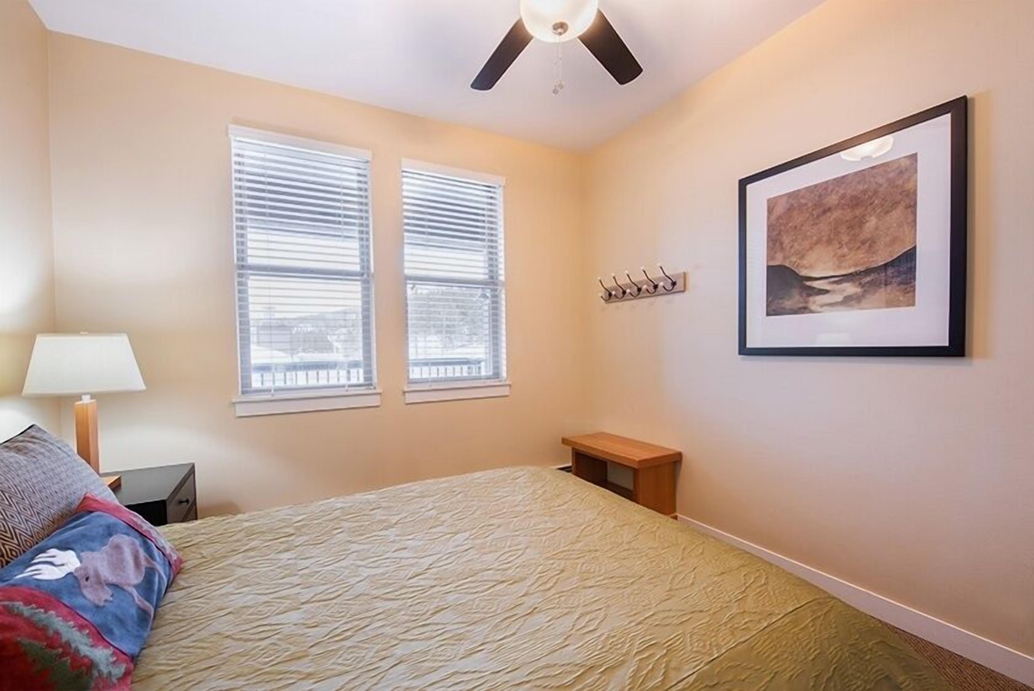 A bedroom with a bed, two windows with blinds, a ceiling fan, a bedside table, a lamp, and a framed picture on the wall.