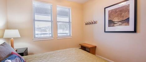 A bedroom with a bed, two windows with blinds, a ceiling fan, a bedside table, a lamp, and a framed picture on the wall.