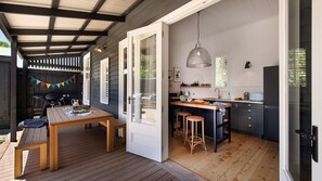 A modern kitchen with a wooden island, a dining area with a table and chairs, and a covered outdoor space with a grill. - The Elm Tree- Designer Home in the heart of Daylesford (Daylesford)
