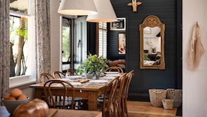 A dining area with a wooden table set for four, surrounded by wooden chairs. There is a large mirror on the wall, a potted plant on the table, and a window with curtains. - The Elm Tree- Designer Home in the heart of Daylesford (Daylesford)