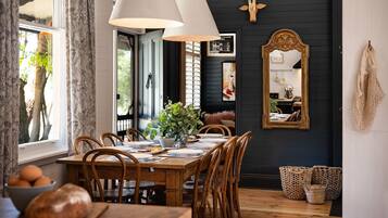A dining area with a wooden table set for four, surrounded by wooden chairs. There is a large mirror on the wall, a potted plant on the table, and a window with curtains.